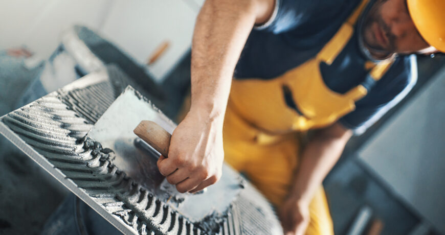 Closeup of a handyman applying adhesive with a trowel material onto a tile before installing it on a bathroom wall. He's wearing yellow uniform and helmet. Tilt shot, selective focus.